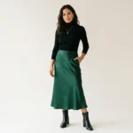 image of a confident modern woman wearing a flowing satin midi skirt with a tucked-in fitted turtleneck top and elegant ankle boots, minimalist fashion aesthetic, soft natural lighting, neutral clean background
