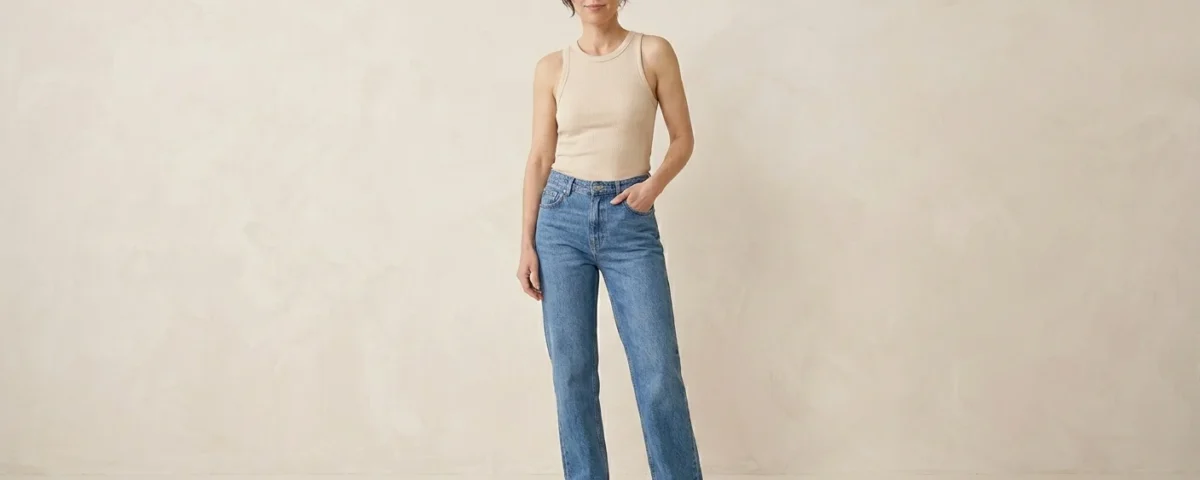 Photorealistic fashion image of a woman standing in a relaxed confident pose wearing classic straight-leg jeans and a simple fitted top. Clean minimal fashion studio background #F5F2ED, soft lighting highlighting the straight silhouette of the jeans, editorial magazine style,