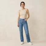 Photorealistic fashion image of a woman standing in a relaxed confident pose wearing classic straight-leg jeans and a simple fitted top. Clean minimal fashion studio background #F5F2ED, soft lighting highlighting the straight silhouette of the jeans, editorial magazine style,