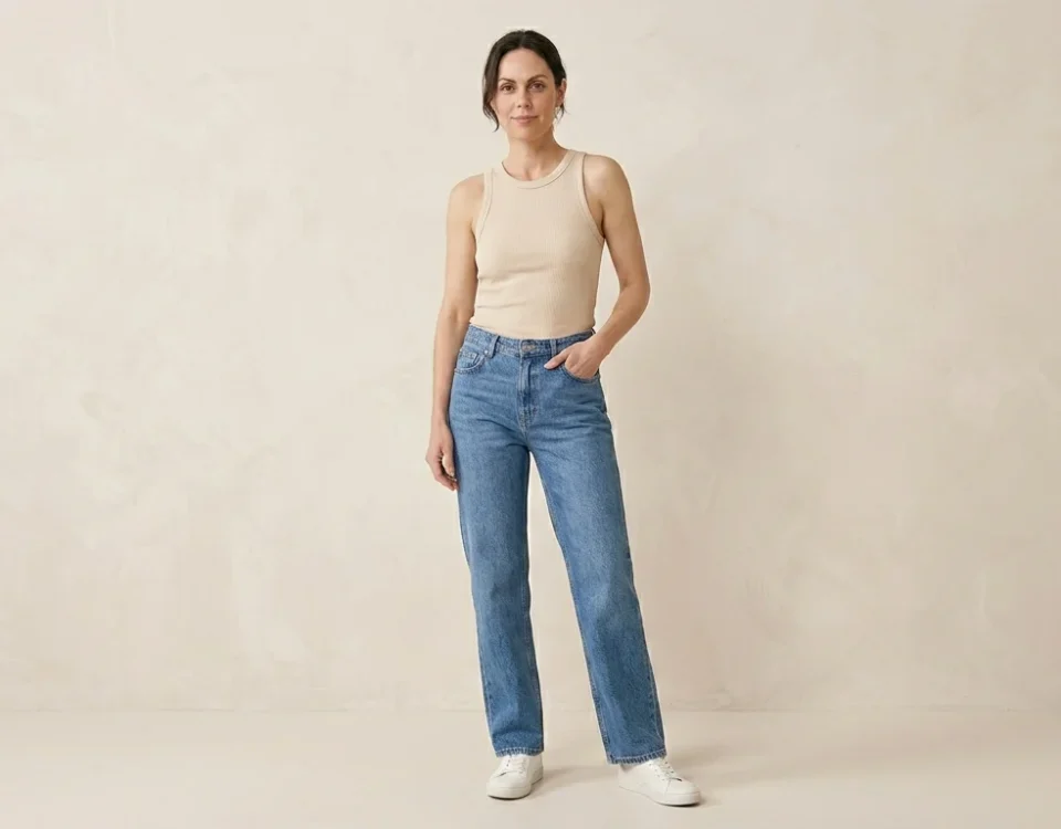 Photorealistic fashion image of a woman standing in a relaxed confident pose wearing classic straight-leg jeans and a simple fitted top. Clean minimal fashion studio background #F5F2ED, soft lighting highlighting the straight silhouette of the jeans, editorial magazine style,